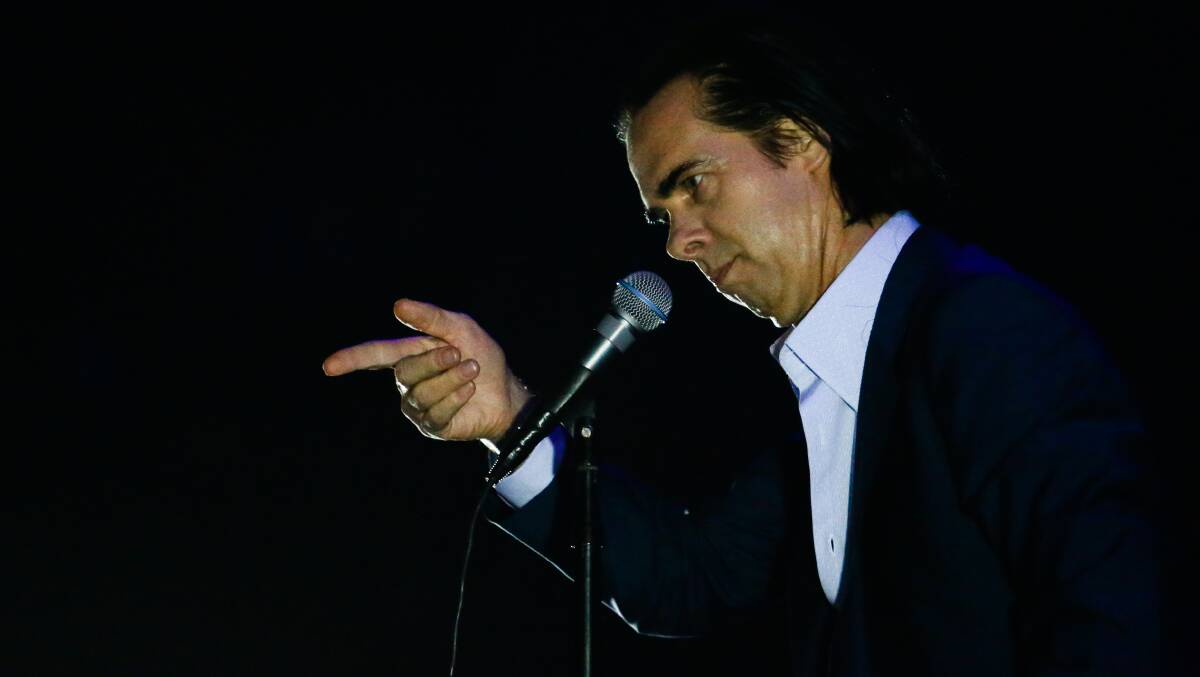 Nick Cave points his finger at the crowd as he leans over a microphone. 